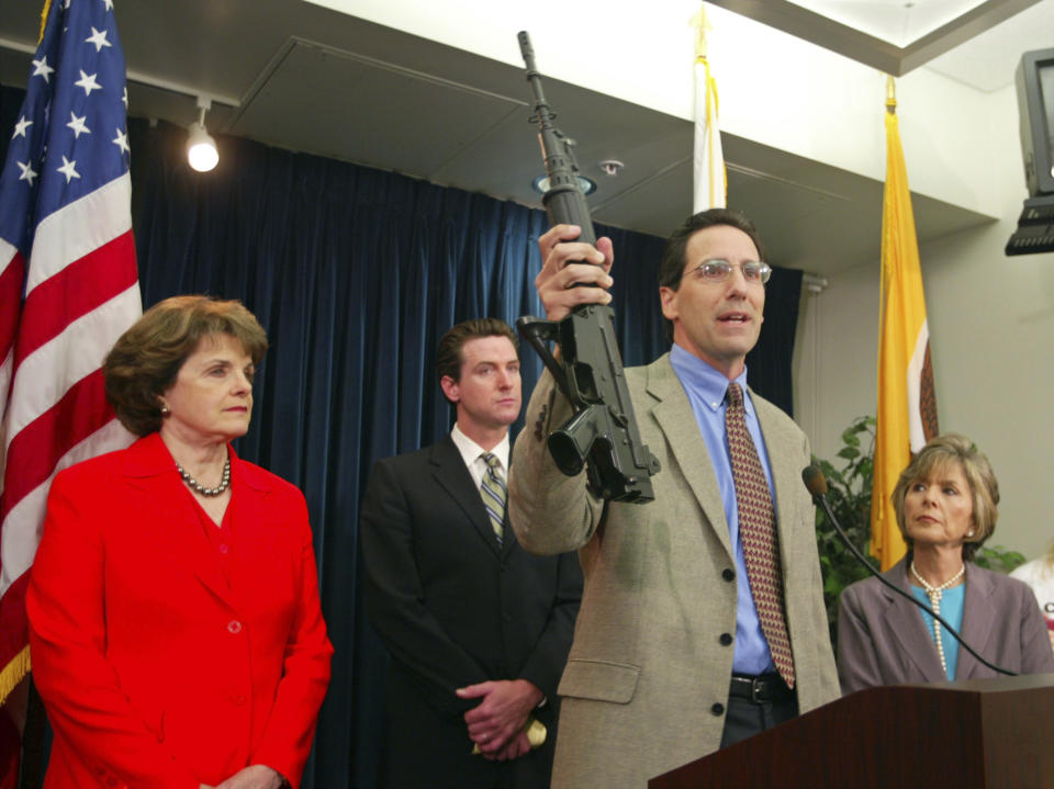 FILE - In this June 29, 2004, file photo, Steve Sposato, who lost his wife in a shooting, holds an automatic rifle as Sen. Dianne Feinstein, D-Calif., left, then San Francisco Mayor Gavin Newsom, second left, and Sen. Barbara Boxer, D-Calif., right, look on at a news conference in San Francisco. Sposato's wife, Jody Sposato, was killed in 1993 in the high-rise shooting at a San Francisco law firm. U.S. District Judge Roger Benitez of San Diego ruled Friday, June 4, 2021, that the state's definition of illegal military-style rifles unlawfully deprives law-abiding Californians of weapons commonly allowed in most other states and by the U.S. Supreme Court. Gov. Newsom condemned the decision, calling it "a direct threat to public safety and the lives of innocent Californians, period." (AP Photo/Eric Risberg, File)