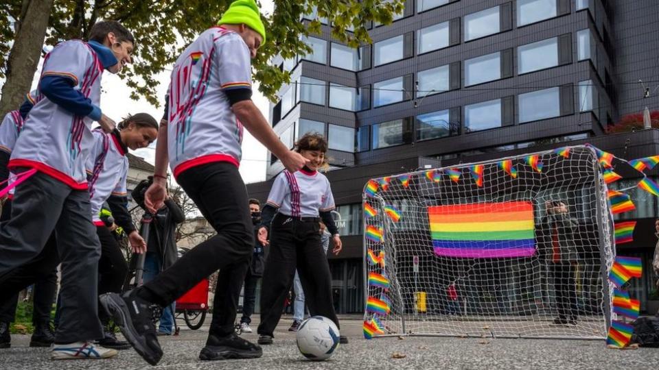 Protesters kick a ball into a goal with rainbow flags on it