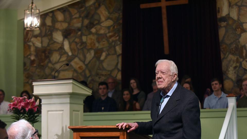 jimmy carter stands next to a wooden podium and smiles as he looks forward, he wears a dark suit jacket and blue collared shirt, people are sitting in front of and behind him