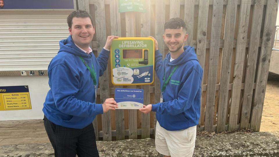 Two men in blue hoodies standing next to a defib.