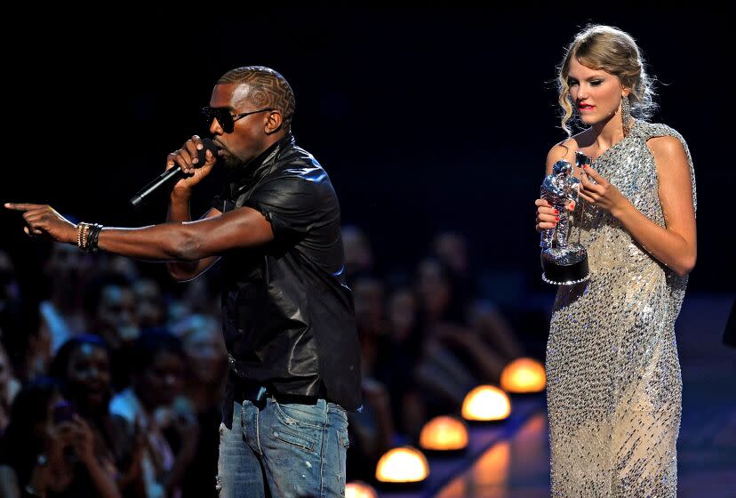 Kanye West in a black shirt, jeans and sunglasses holding a microphone on stage with Taylor Swift holding an award