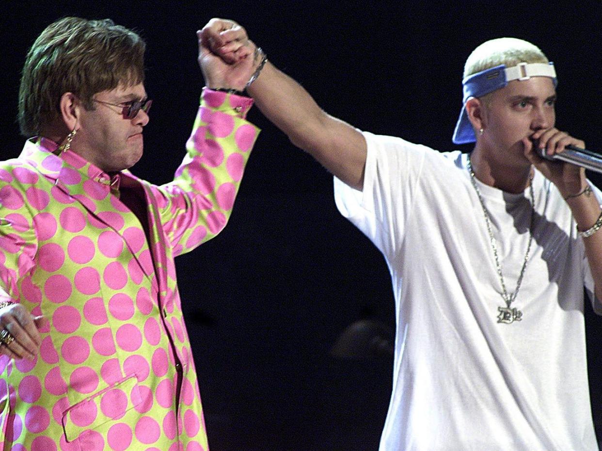 Elton John in a yellow suit jacket with large pink circles holding his hand up to hold Eminem's hand, whose wearing a white t-shirt, backwards hat, and a speaking into a microphone.