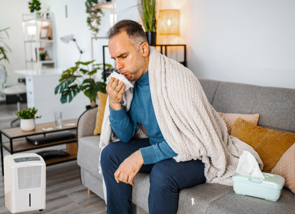 Man with a blanket around his shoulders, sitting on the couch, sneezing into a tissue.