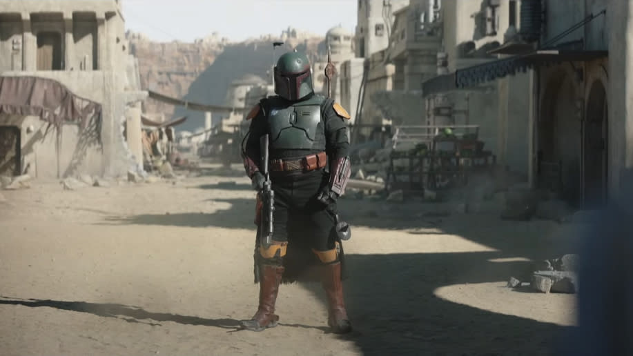 Boba Fett in armor stands in a dusty street with buildings in the background