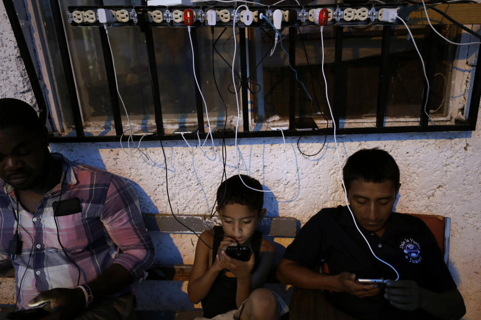 In this July 30, 2019, photo, migrants from Africa and Latin America check their phones among cables of charging phones plugged into sockets at El Buen Pastor shelter for migrants in Cuidad Juarez, Mexico. (AP Photo/Gregory Bull)