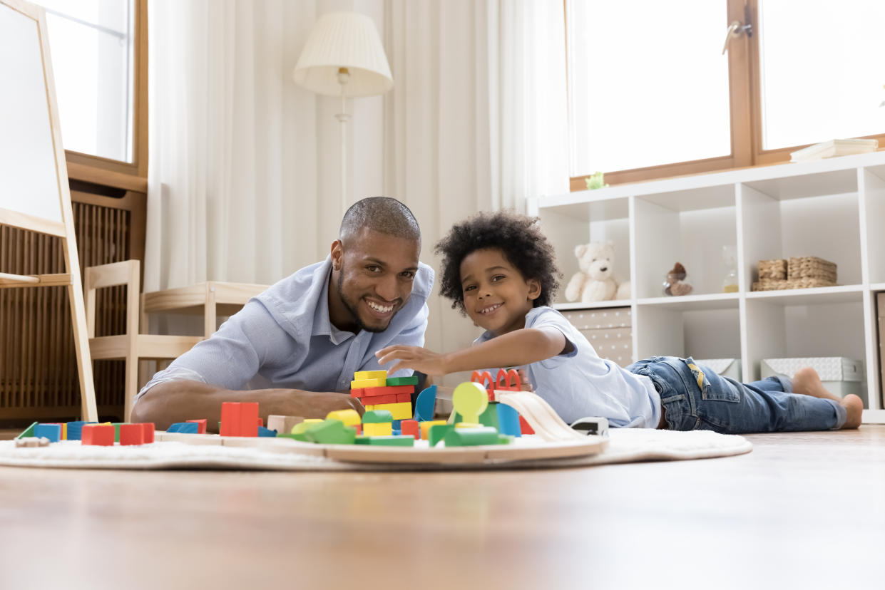 Portrait of happy bonding young african american father and small adorable kid son lying on floor carpet, enjoying playing wooden toys, posing having fun on weekend together at home, looking at camera