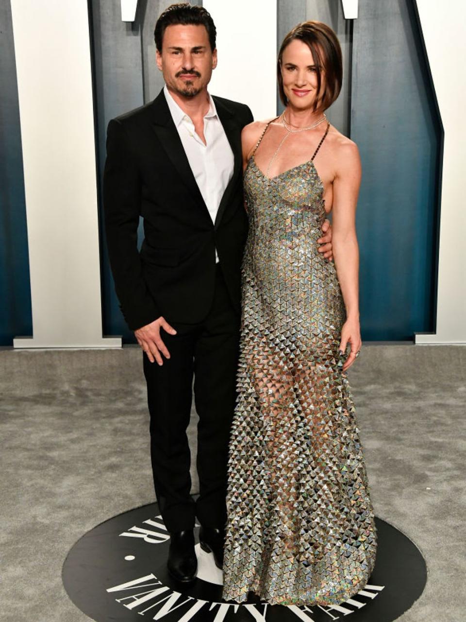 Juliette Lewis and Brad Wilk attending the 2020 Vanity Fair Oscar Party. She is wearing a gold spaghetti strap evening gown while he is in a black suit with a white shirt. 
