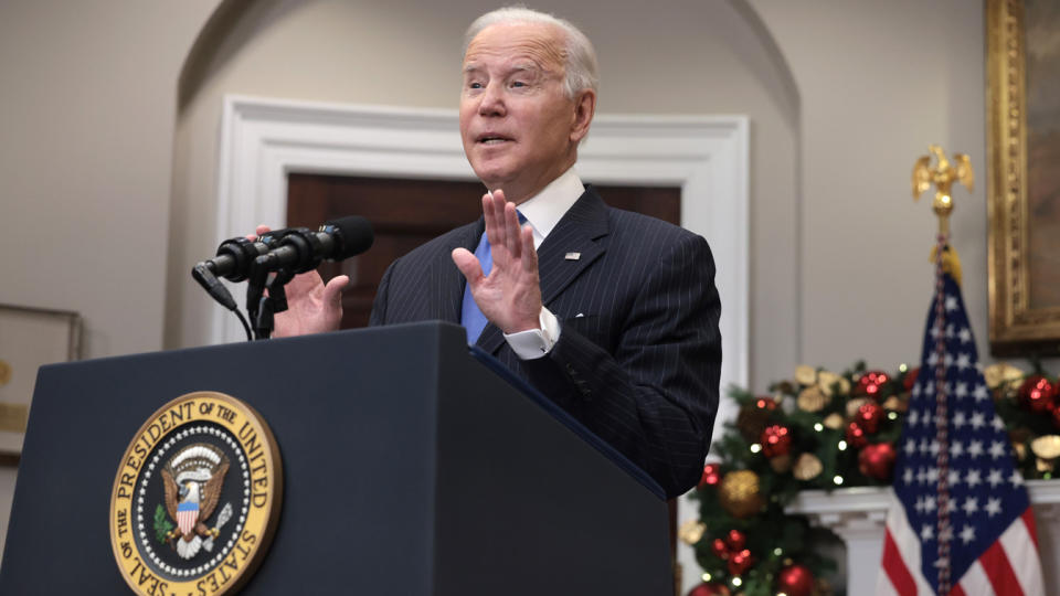 President Biden stands with palms raised at a podium that bears a presidential seal, in front of an American flag and holiday decorations.