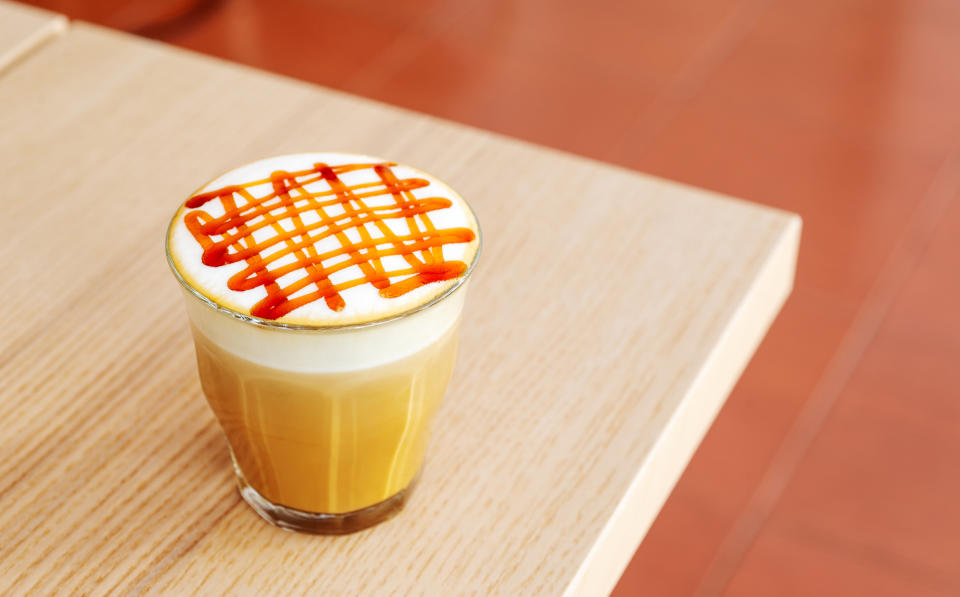 A glass of latte topped with foam and caramel drizzle sits on a light wooden table. No people are present in the image