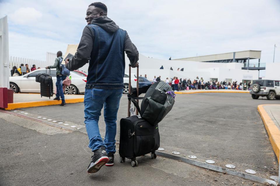 A man, in a hooded shirt, vest and jeans, is seen from behind wheeling his suitcase with a carryall slung over the handles