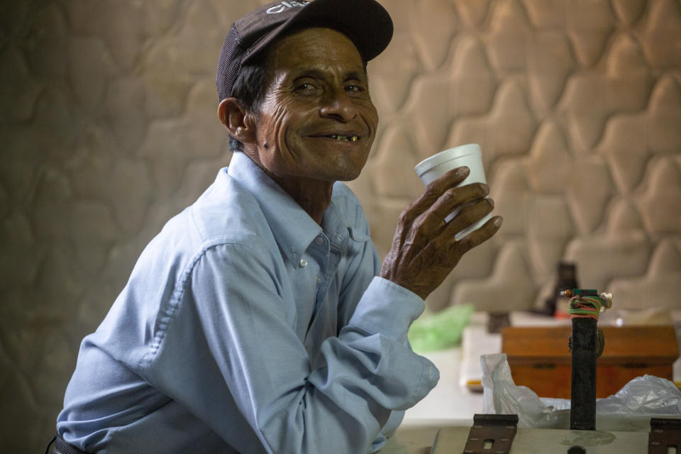 El vendedor de helados Cornelio Tepen, de 59 años, sonríe a la cámara mientras bebe café después de llegar a la casa destartalada que solía almacenar los carros de los vendedores y donde algunos de ellos viven durante la pandemia de COVID-19 en la Ciudad de Guatemala, el martes 26 de mayo de 2020. (AP Foto/Moises Castillo)