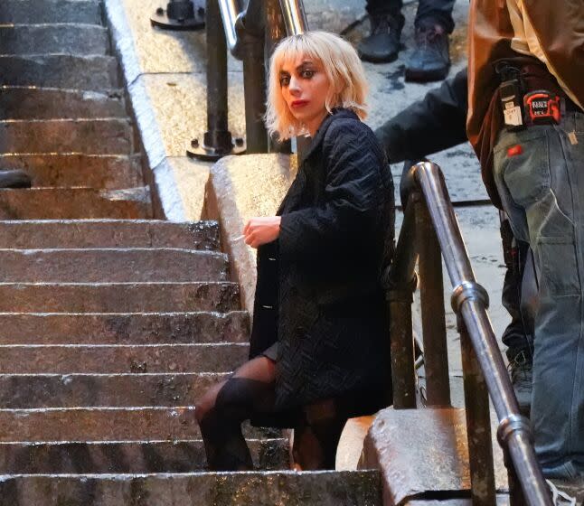 Lady Gaga dressed as Harley Quinn with white makeup and a dark coat leans against a railing along set of concrete stairs