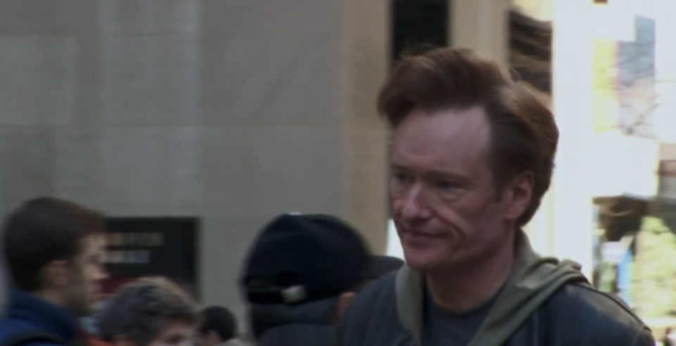 Conan O'Brien in a casual outfit with a backpack, amidst a crowd outside