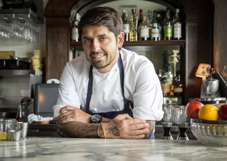 A smiling man in chef's whites and a blue apron leans on a marble counter.