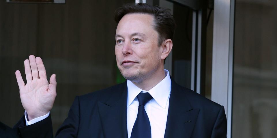 Elon Musk, wearing a black suit and tie, waves to photographers outside a courthouse