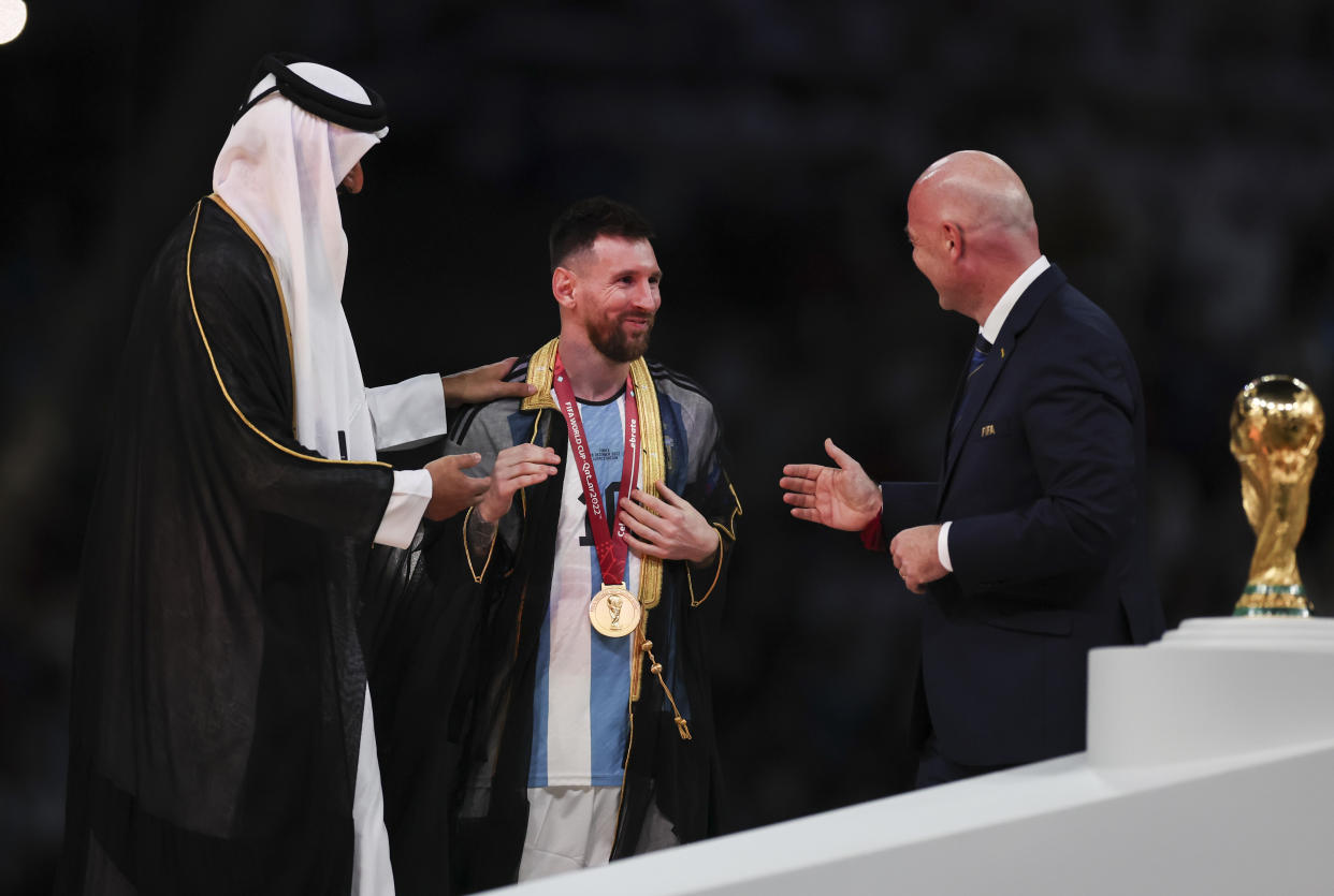 LUSAIL CITY, QATAR - DECEMBER 18:  Lionel Messi of Argentina is presented a traditional black bisht robe by Sheikh Tamim bin Hamad Al Thani, Emir of Qatar, while Gianni Infantino, President of FIFA, looks on after the FIFA World Cup Qatar 2022 Final match between Argentina and France at Lusail Stadium on December 18, 2022 in Lusail City, Qatar. (Photo by Ian MacNicol/Getty Images)