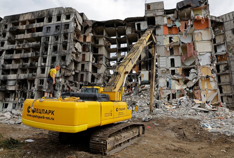 A worker stands on an excavator in front of a destroyed apartment building in Mariupol