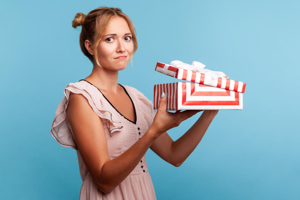 Portrait of upset young adult blonde female with bundles, holding red striped present box, has sad expression, being disappointed with birthday gift. Indoor studio shot isolated on blue background.