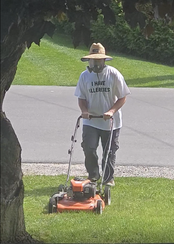 Person mowing the lawn wearing a wide-brimmed hat, a mask, and a shirt that reads "I Have Allergies." Outdoor scene with greenery in the background