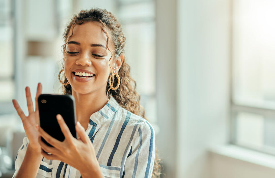 A woman smiles and waves at her phone inside a bright, modern room, engaging in a video call. She wears hoop earrings and a striped shirt