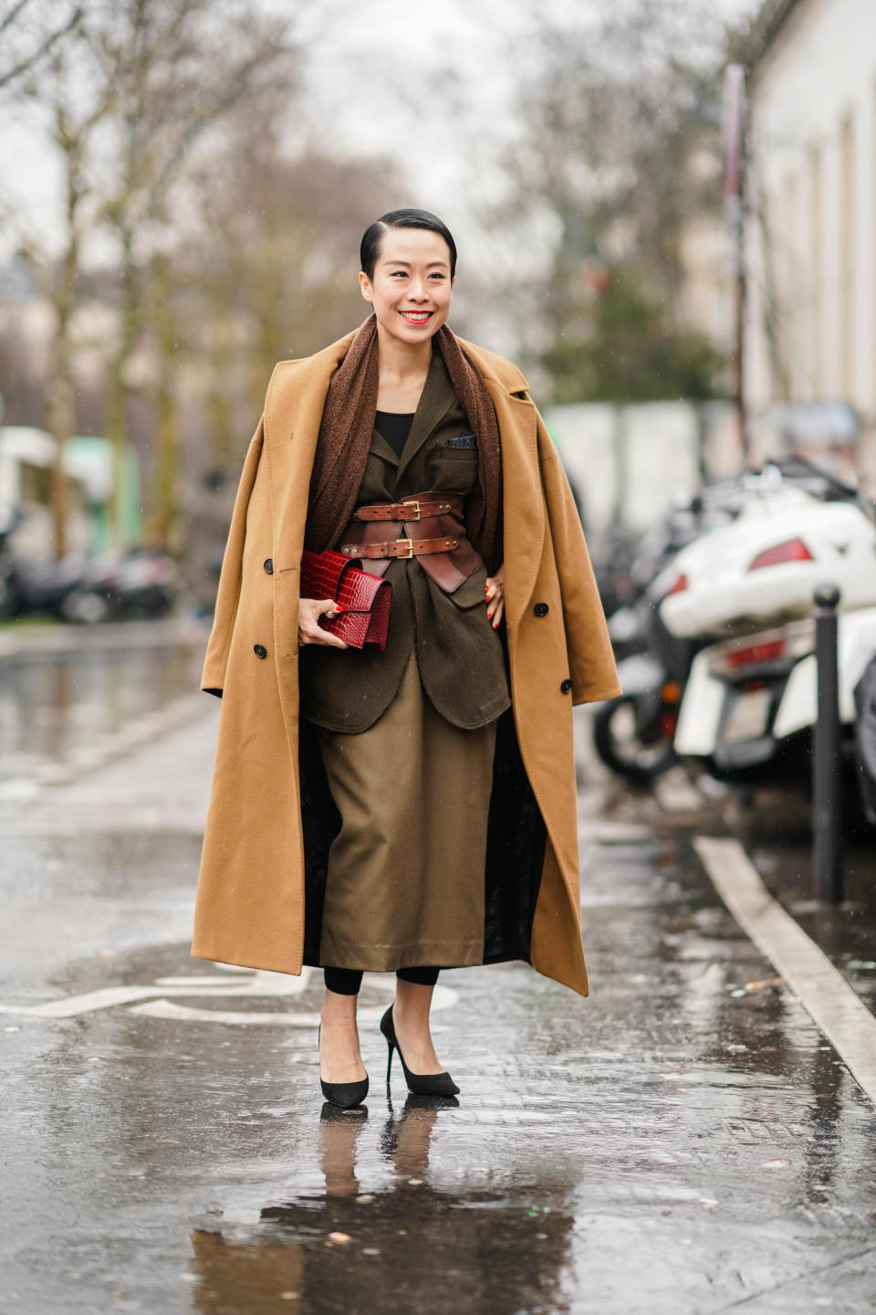 guest wears a long camel coat, a brown scarf, a red clutch, a khaki jacket, a yellow bronze long skirt, black leggings, black stilettos, a red double buckle obi belt.