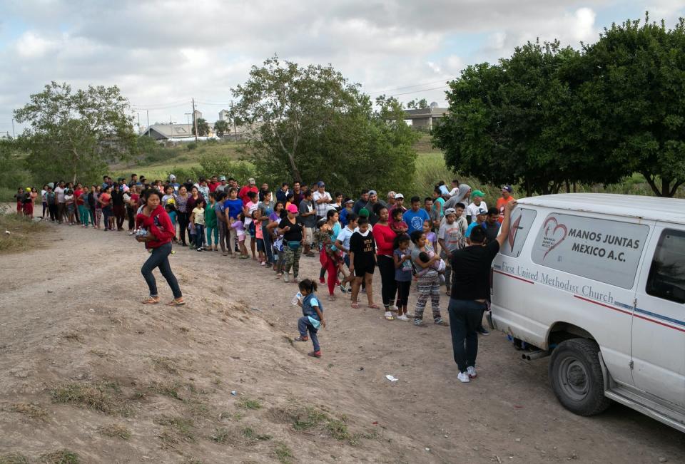 Line of people standing behind a van, with children playing in dirt in foreground