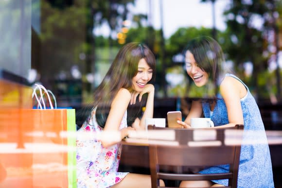 Two young Asian women in a cafe, laughing over mobile device.