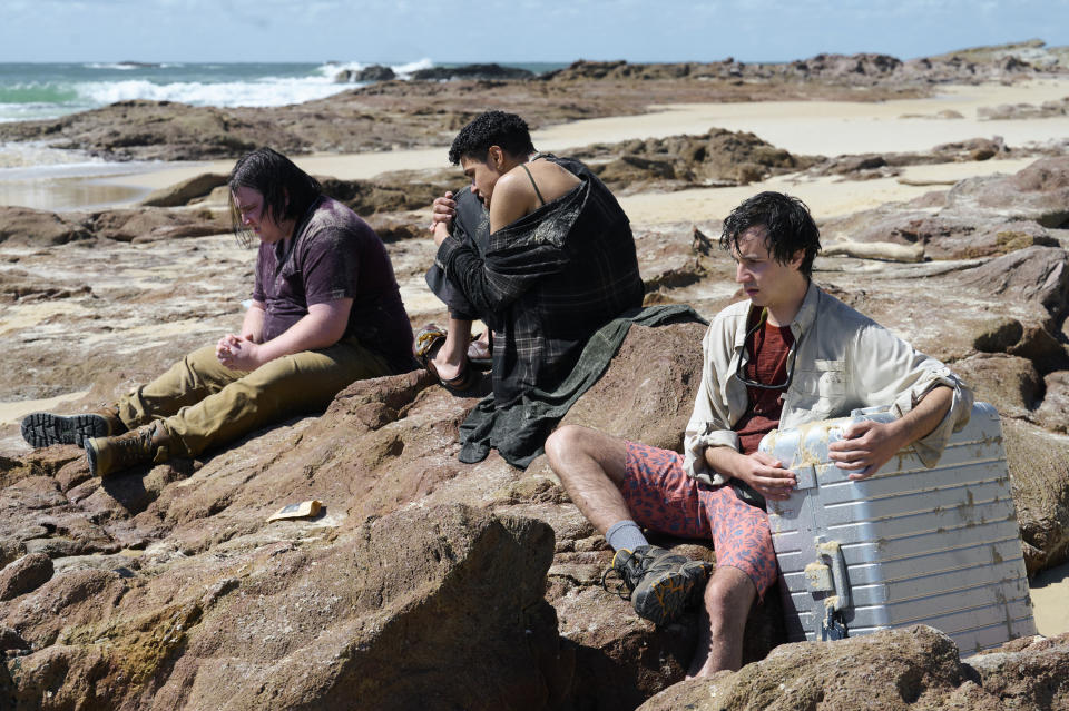 A still from the series The Wilds showing characters sitting on sandy rocks by the ocean