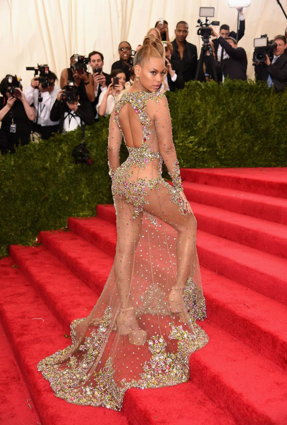 Beyonce poses while walking up the steps of the red carpet at the Met Gala, wearing a sheer dress with colorful jewels on the back and hem.