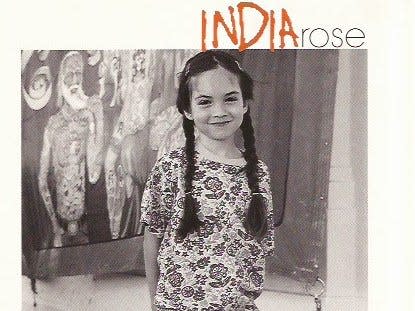 A black and white photo of India Kushner in front of a mural of a man with a tattoo. She has dark hair in braids and dark eyes. She smiles slightly and wears a floral t-shirt. 
