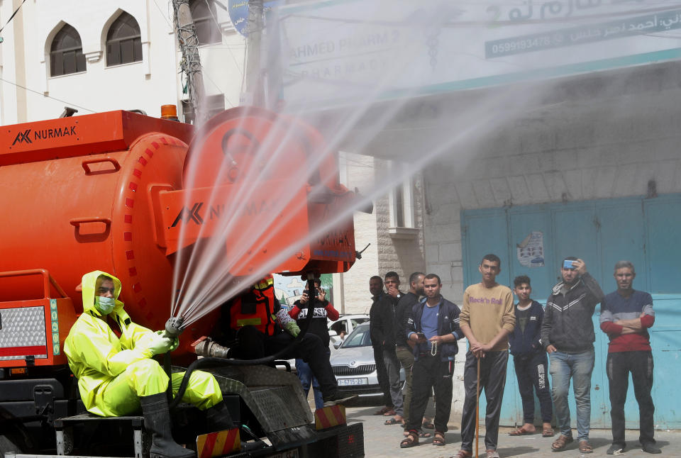 Residents watch workers wearing protective gear spray disinfectant as a precaution against the coronavirus, at the main road of closed market of Shijaiyah neighborhood in Gaza City, Friday, March 27, 2020. Gaza municipality close all the weekly Friday markets in Gaza starting from today. (AP Photo/Adel Hana)