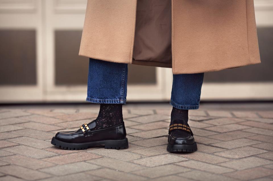 person wearing long tan coat, dark jeans, black socks, and black loafers