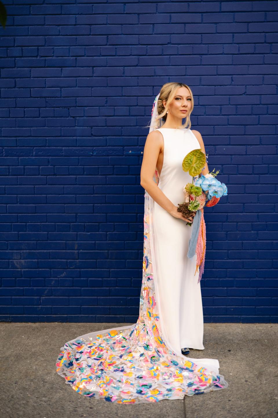 A bride in a white dress and rainbow veil holds a bouquet in front of a blue, brick wall.