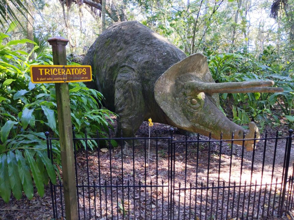 A statue of a triceratops dinosaur next to lush green trees, behind a fence.