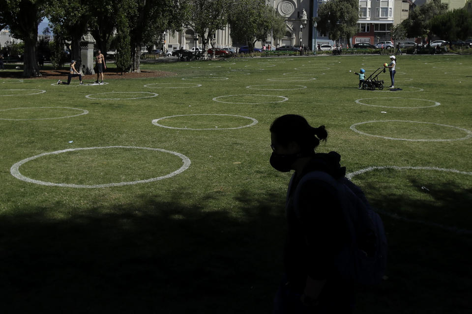 FILE - In this May 23, 2020, file photo a woman wears a face mask while walking in front of circles designed to help prevent the spread of the coronavirus by encouraging social distancing at Washington Square park in San Francisco. As some parts of the nation continue to ease stay-at-home orders meant to slow the spread of the coronavirus, the economy remains in free fall. President Donald Trump says that the country is anxious to get back to work and that pent-up consumer demand can turn things around quickly. Presumptive Democratic nominee Joe Biden counters by urging caution and heeding medical and scientific experts. (AP Photo/Jeff Chiu, File)