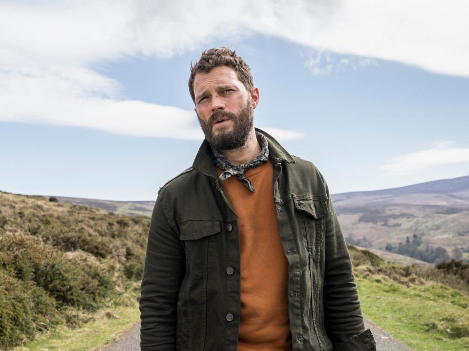 jamie dornan in the tourist, wearing an orange shirt and green jacket, standing in a remote road