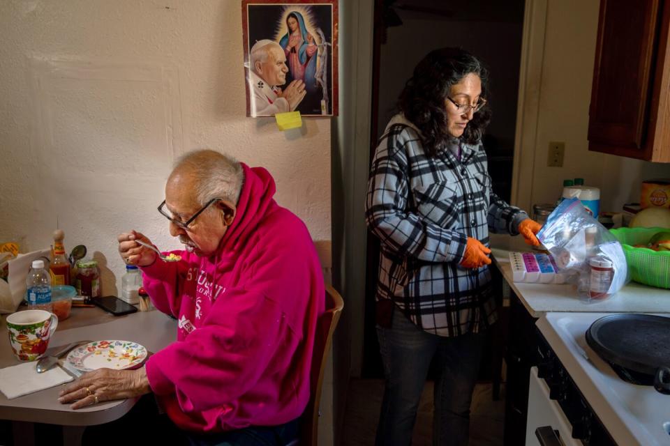 A woman stands at a kitchen counter refilling medication tray and a man sits at a kitchen table eating.