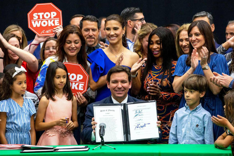 Gov. Ron DeSantis, beaming, holds up a folder containing a signed document, HB 7, 