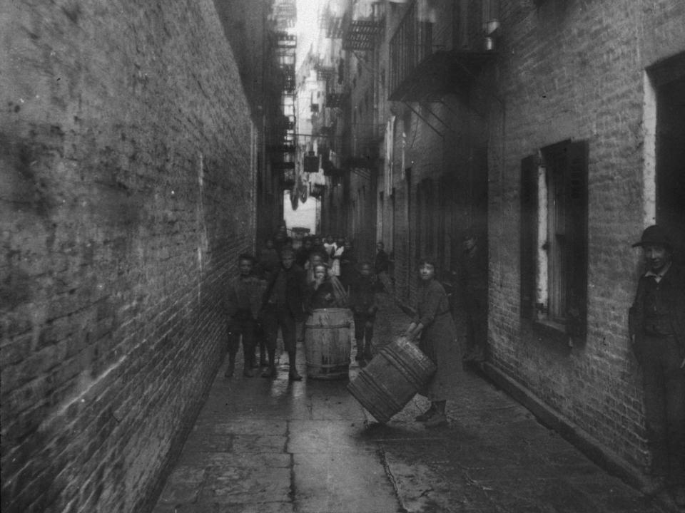 Children play with barrels in an alley between tenement buildings in Gotham Court in New York City.