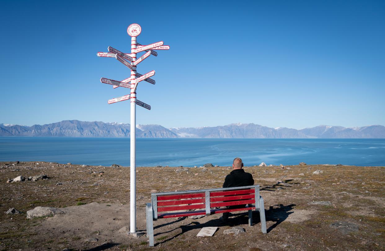 Someone sits on a red bench next to a white post with signs radiating out from it showing the distances to various cities and appears to look out over blue water and mountains beyond.
