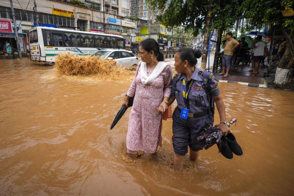 Women wade through the muddy brown waters on a flooded street, near buses and storefronts