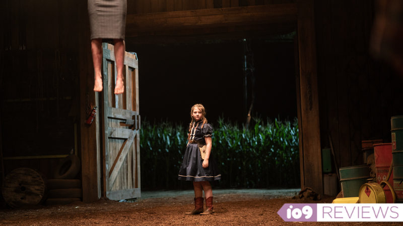 A small girl stands in a barn with a cornfield behind her and a body hanging next to her