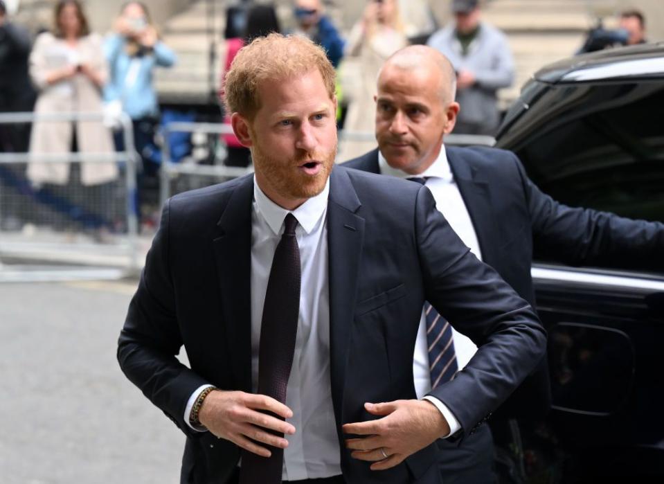 prince harry, wearing a black suit and tie, straightens his jacket and looks to the side as he walks away from a black car outside, with another man standing behind him