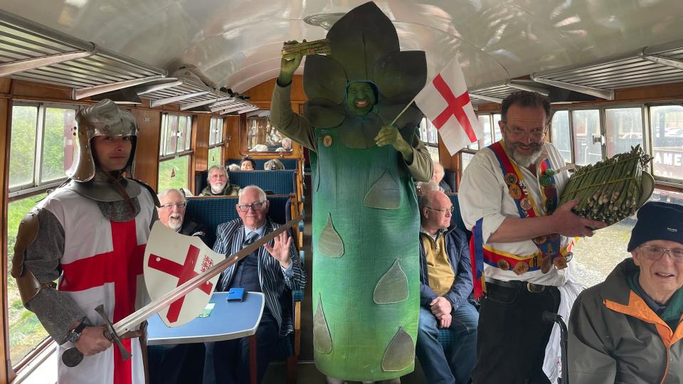 A man dressed as a giant asparagus on a train with people dressed as St George