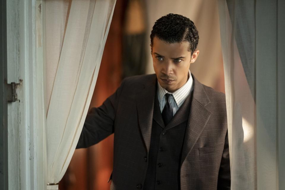 Jacob Anderson wearing a classic brown suit and tie, stands in a doorway with white curtains in a scene from "Interview with the Vampire"