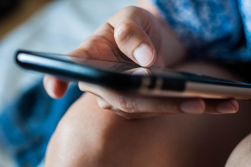 A close-up of a person's hand holding a smartphone, touching the screen with their thumb. The background is blurred