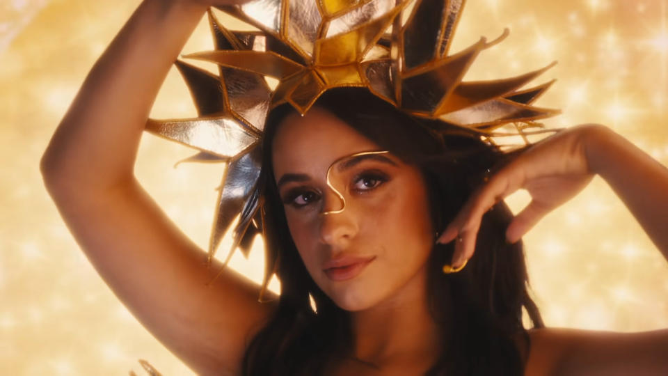 Headshot of Camilla with her hands raised and wearing a dramatic headdress