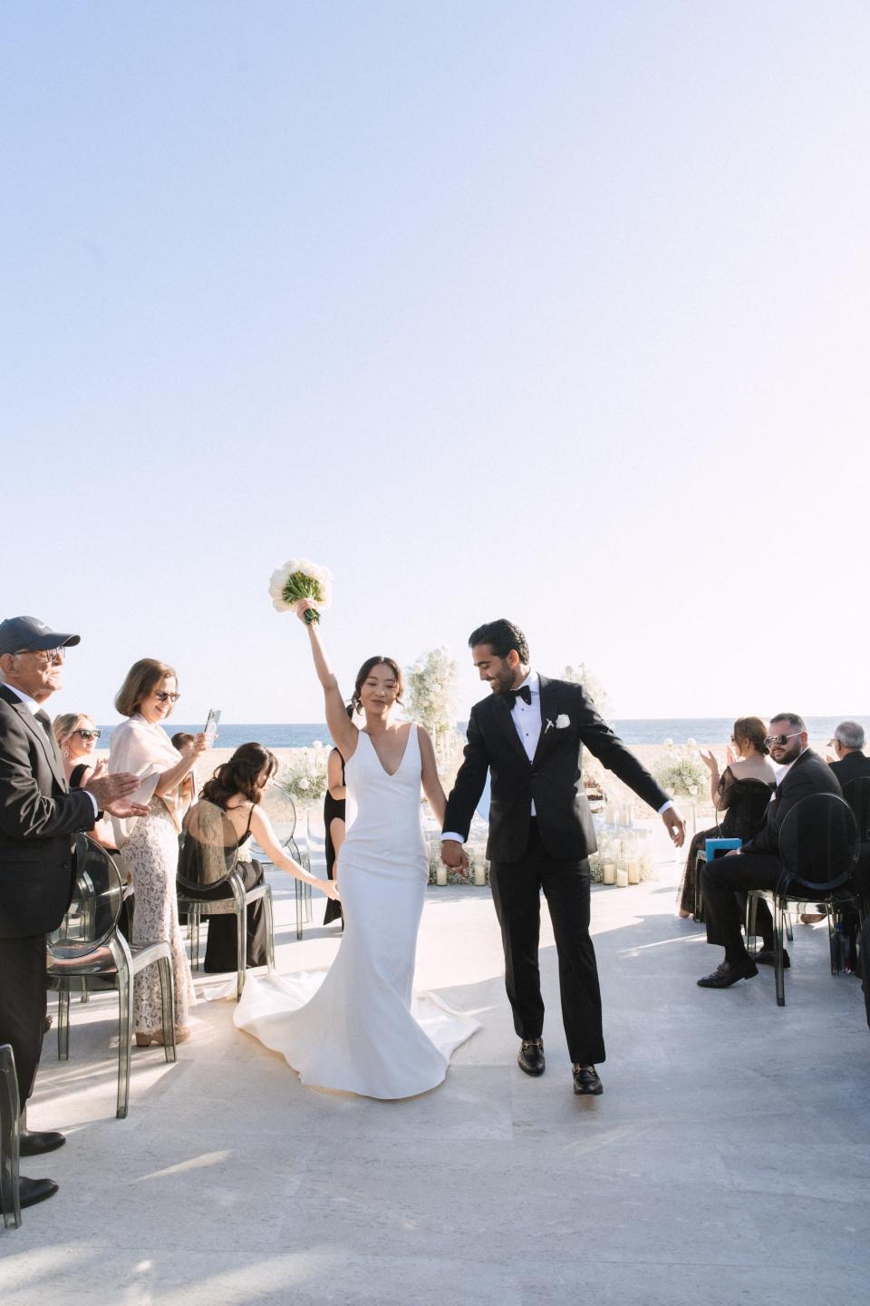 A bride and groom exit their wedding ceremony on a beach. The bride raises her hand holding her bouquet in excitement.