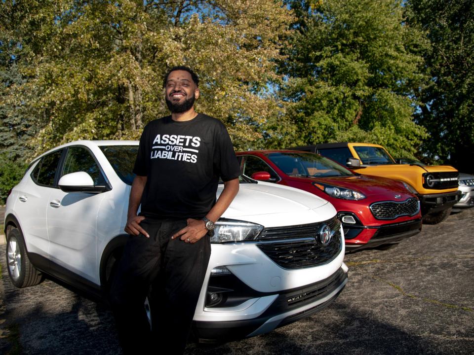 Lavell Riddle stands in a parking lot in front of three cars. one car is white, one is red, and one is yellow