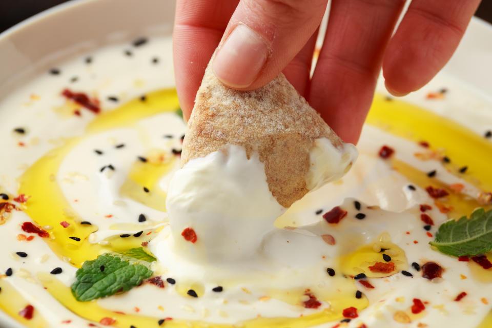 A hand dips a piece of pita bread into a bowl of yogurt sauce garnished with olive oil, mint leaves, black sesame seeds, and red pepper flakes